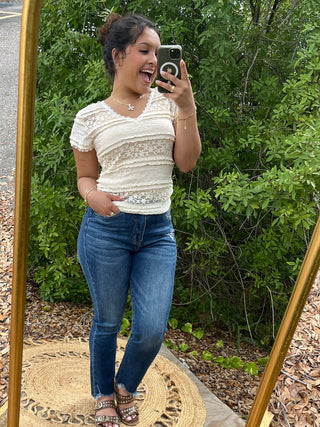 Cream lace short-sleeve top with striped texture and sheer panels, paired with dark blue jeans.
