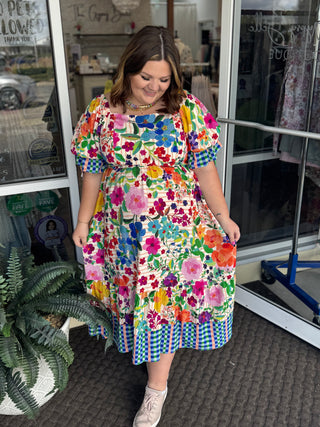 Colorful floral print dress with bubble sleeves and blue checkered border hem on white background.
