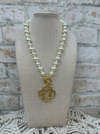 Cream pearl necklace with gold toggle clasp and ornate gold clover charm pendant on display bust.
