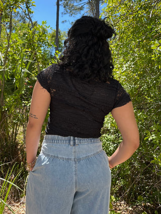 Black lace short-sleeve top with textured pattern and sheer details, worn with light-wash denim pants.
