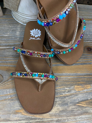 Tan thong sandals with multicolored rhinestone straps and crystal embellishments, featuring a daisy logo.
