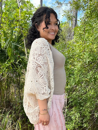 Cream crochet shrug with open lace pattern worn over gray tank top and pink pants.
