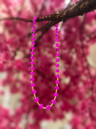 Hot pink glass bead necklace with gold spacers hanging from branch against blurred pink floral background.
