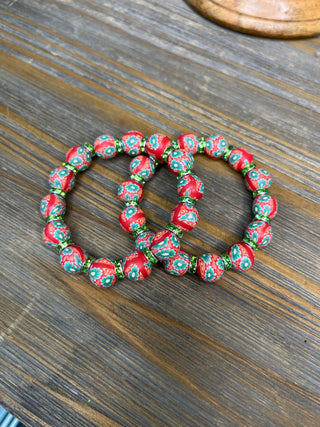 Red floral clay bead stretch bracelets with green and gold accents on wooden surface.
