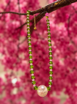 Green faceted beads and pearl pendant necklace against pink floral background.
