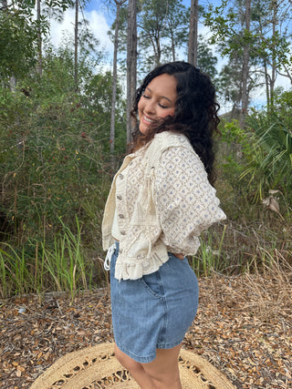 Cream floral button-up jacket with delicate lace details and ruffled hem over denim shorts.
