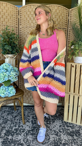 Oversized cream cardigan with multicolor horizontal stripes in pink, blue, orange and gray worn over bright pink tank top.
