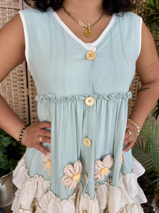 Light blue sleeveless dress with white trim, wooden button details, ruched waist, and embroidered daisy hem on cream ruffle s
