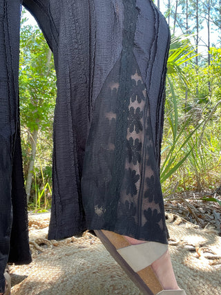 Black wide-leg pants with geometric mesh panel detail worn on sandy beach with tropical foliage.
