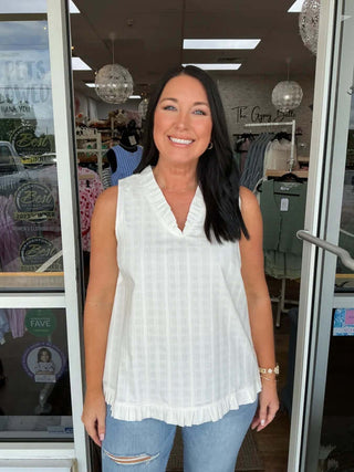 Alt text: "White Textured Cotton V Neck Sleeveless Top by Doe & Rae, displayed on hanger against a white brick wall.
