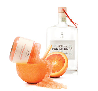 Blood orange glowrita body polish jar with fresh orange slices and tequila bottle displayed together.
