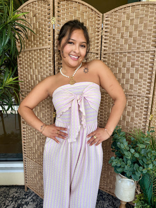 Lavender striped strapless jumpsuit with bow detail at chest, worn with pearl necklace.
