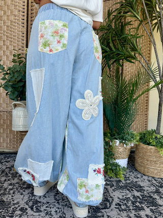 Light blue denim joggers with floral patchwork pockets, white crochet flower appliqué, and distressed details.
