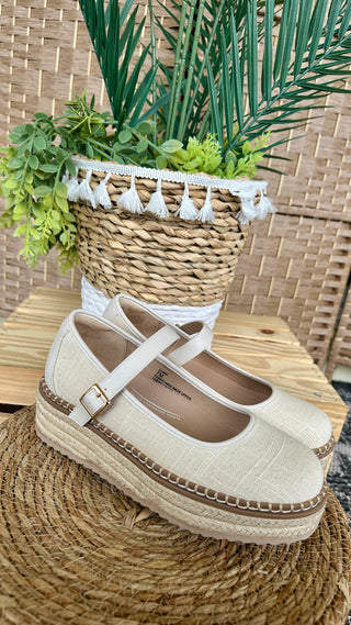 Cream linen espadrille Mary Jane flat with white ankle strap and jute sole on woven placemat.
