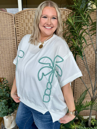 White oversized tee with teal palm tree graphic design, worn with jeans.
