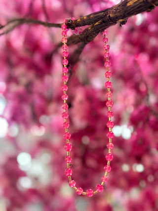Pink glass bead necklace with gold spacers hanging from branch with blurred pink flowers background.
