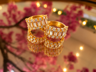 Gold huggie hoop earrings with baguette and round crystal stones, displayed with pink flowers and warm lighting.
