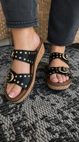 Black wedge sandals with studded straps, gold buckles, and cork soles on model wearing dark denim.
