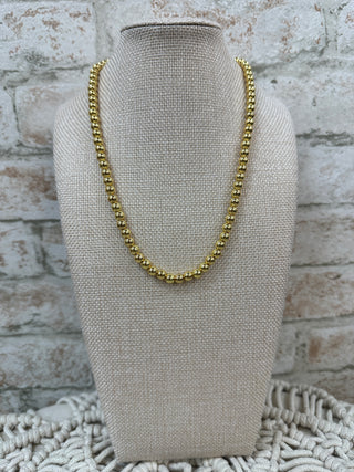 Gold beaded collar necklace on cream linen display bust against whitewashed brick background.
