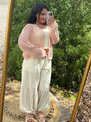 Pink crochet cardigan with open weave pattern worn over white top and cream pants.
