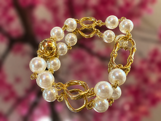 Gold-toned bracelet with white pearl beads and ornate gold bamboo-link connectors on pink floral background.
