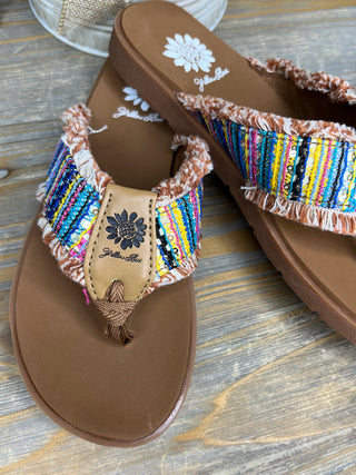 Tan flip flop sandals with colorful striped fringe trim and tassel detail on thong strap.
