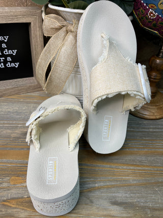 Cream flatform slide sandals with textured linen upper and frayed edges, displayed on wooden surface.
