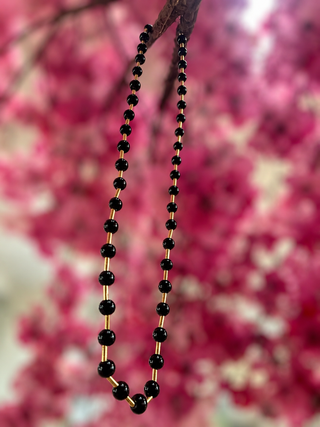 Black beaded necklace with alternating black glass and gold-tone spacer beads against pink floral background.
