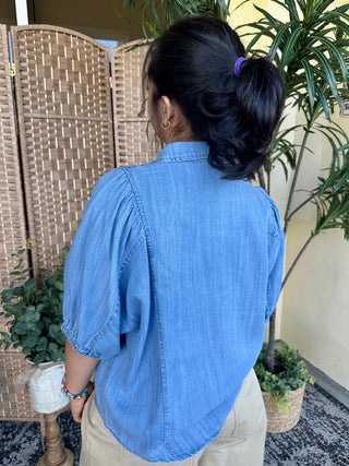 Light blue chambray button-up shirt with puffed short sleeves, worn over cream pants.
