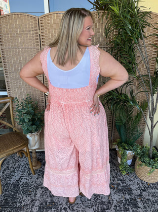 Pink eyelet sleeveless jumpsuit with lace trim and wide-leg cropped silhouette over light blue tank top.

