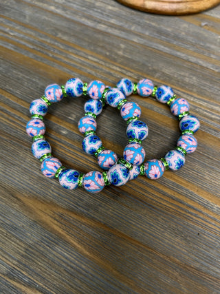 Blue and pink floral clay bead stretch bracelets with green spacer beads on wooden surface.
