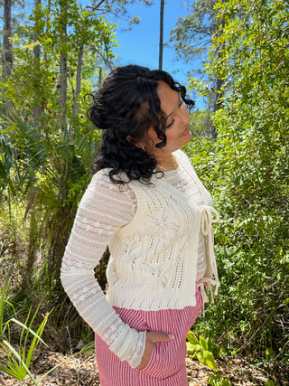 Cream cable knit vest top with lace sleeves and tie waist detail worn with red striped pants.
