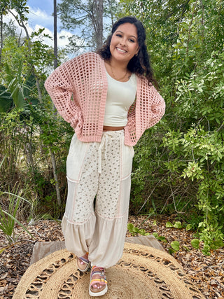 Pink open-weave crochet cardigan layered over white crop top with cream floral pants.
