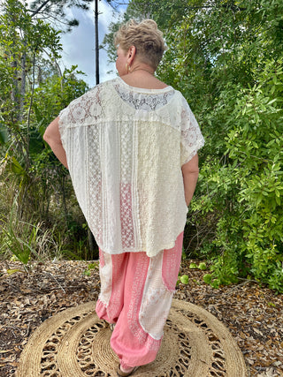 Ivory lace short-sleeve top with geometric pattern and sheer fabric, worn over pink patterned pants.
