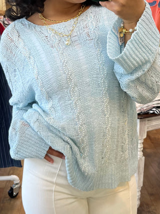 Light blue cable knit sweater with twisted weave texture and long sleeves worn with white pants.
