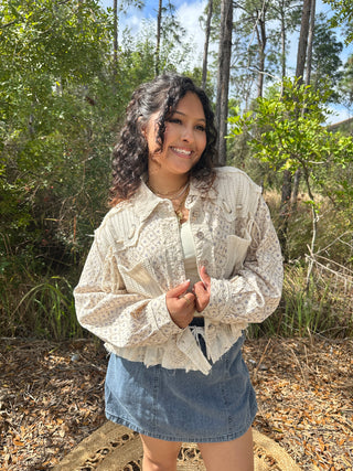 Ivory floral lace jacket with textured fabric and ruffled hem, worn over denim skirt.
