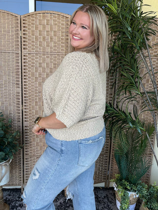 Taupe asymmetrical knit sweater with textured weave and three-quarter sleeves, paired with light wash distressed jeans.
