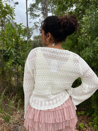 Off-white crochet cardigan with openwork pattern and ruffled hem worn over pink skirt.
