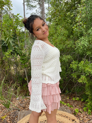 Off-white crochet cardigan with openwork pattern and bell sleeves over pink pleated skirt.
