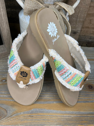 Beige flip-flop sandals with colorful striped fabric cuffs, white fringe trim, and decorative sun emblem detail.
