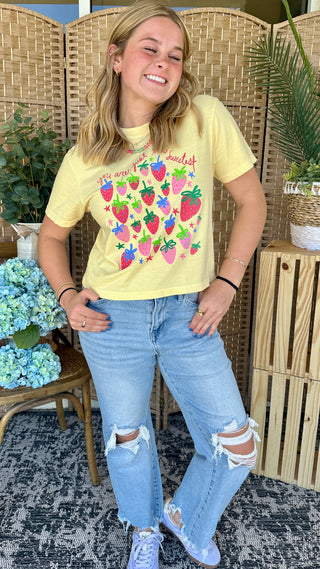Pale yellow crop tee with colorful strawberry and text graphic, styled with distressed blue jeans.
