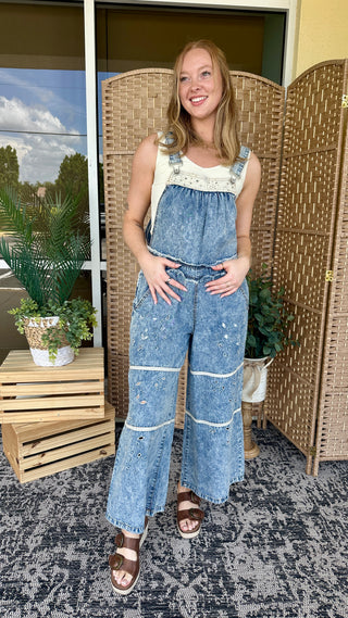 Blue denim overalls with ivory eyelet trim and white stitching detail on front straps and legs.
