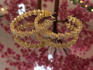 Gold glitter textured hoop earrings with ruffled edge detail displayed against blurred floral background.
