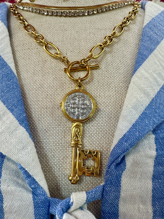 Gold chain necklace with religious medal and key pendant on striped blue and white top.
