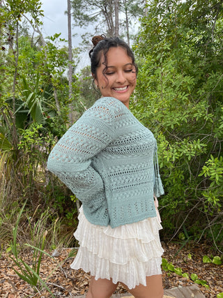 Sage green open-knit cardigan with openwork pattern and tassel trim over cream pleated skirt.
