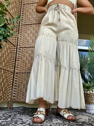 Cream tiered patchwork pants with drawstring waist and textured fabric panels, styled with white leather sandals.
