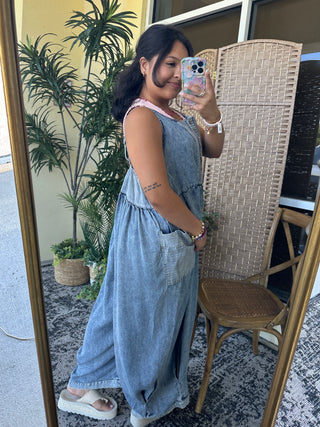 Blue denim sleeveless jumpsuit with large pockets and relaxed fit, styled with white slides.
