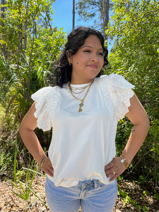 White eyelet top with flutter sleeves and cream trim, paired with light blue jeans.

