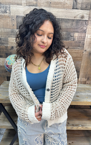 Cream crochet cardigan with ivory trim over blue fitted top, styled with striped pants.
