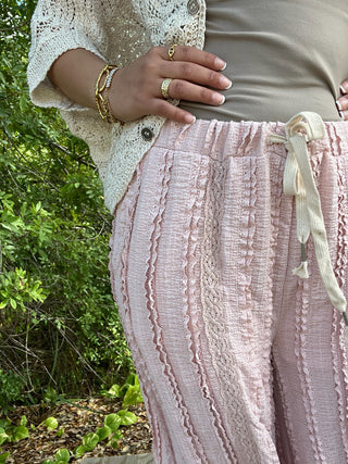 Mauve wide-leg pants with vertical lace and ruffle stripe pattern, drawstring waist, styled with cream lace cardigan.
